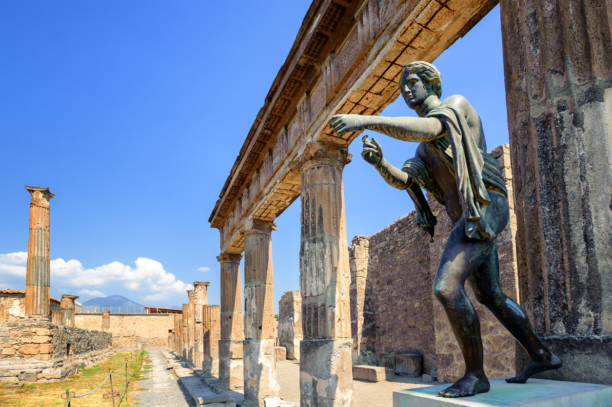 Bronze statue among ancient Roman columns in Pompeii on a sunny day, Mount Vesuvius in the distance